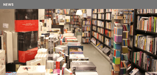 Ein Blick in die Buchhandlung ALEX vom vorderen Ende des großen Tisches, zu sehen sind Stapel mit Büchern und rechts das lange Bücherregal mit Kunst-, Architektur-, Psychologie- und Philosophiebüchern